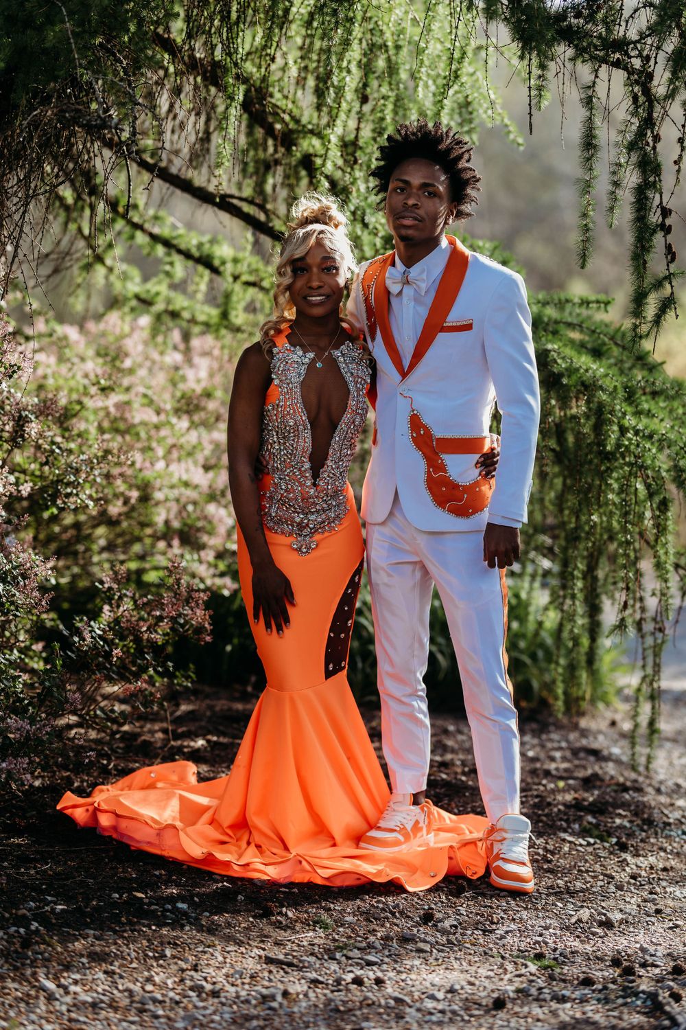 Prom couple posing outdoors in coordinating orange and white formal attire under moss-draped trees.
