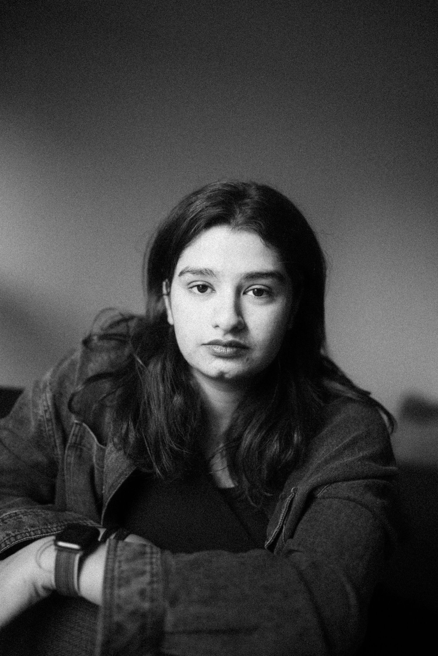 A black and white portrait in moody lighting shows someone wearing a dark jacket and resting their head on crossed arms.