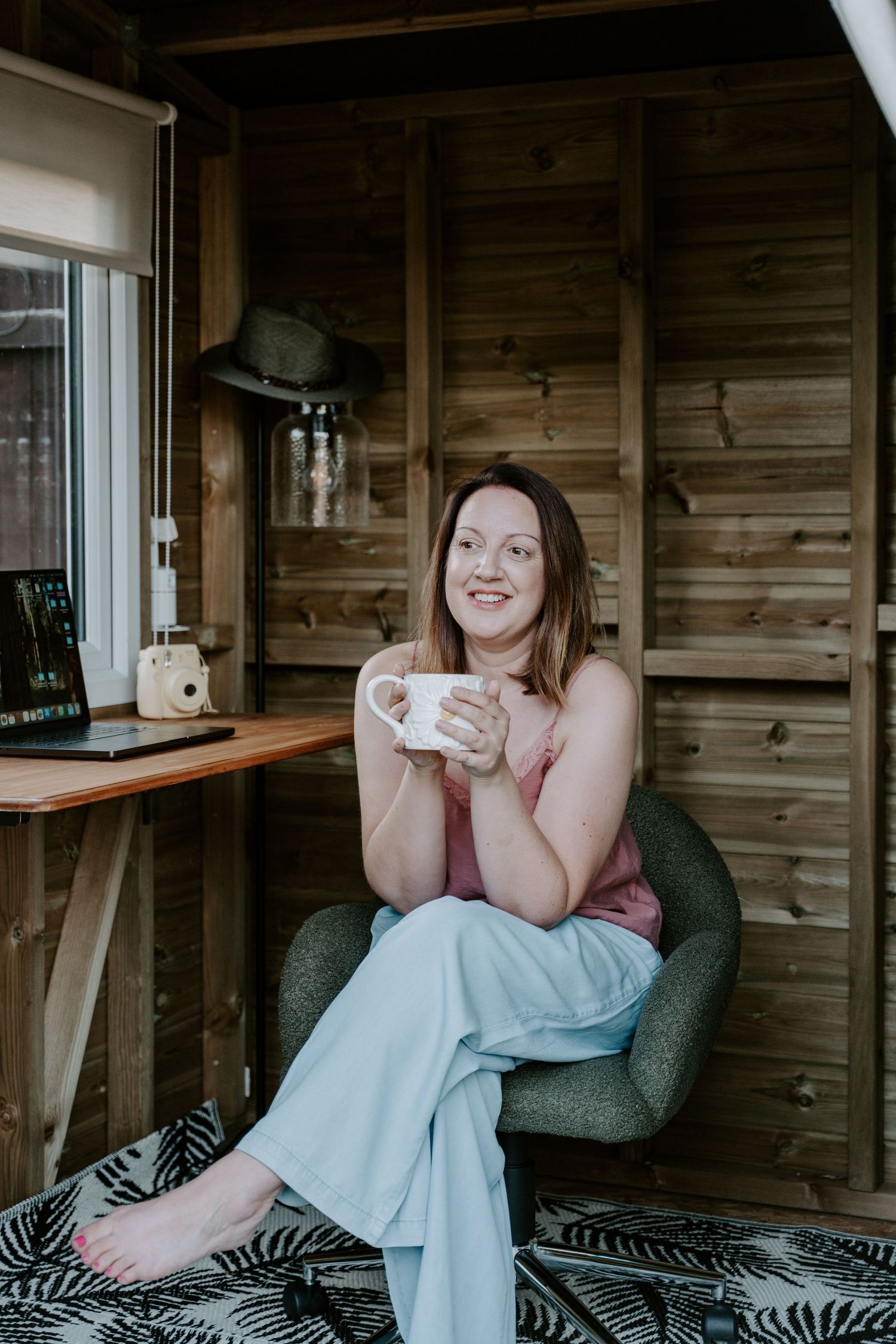 A photographer sat at her desk looking away from camera and holding a mug of tea. She is smiling and is sitting with her legs crossed.