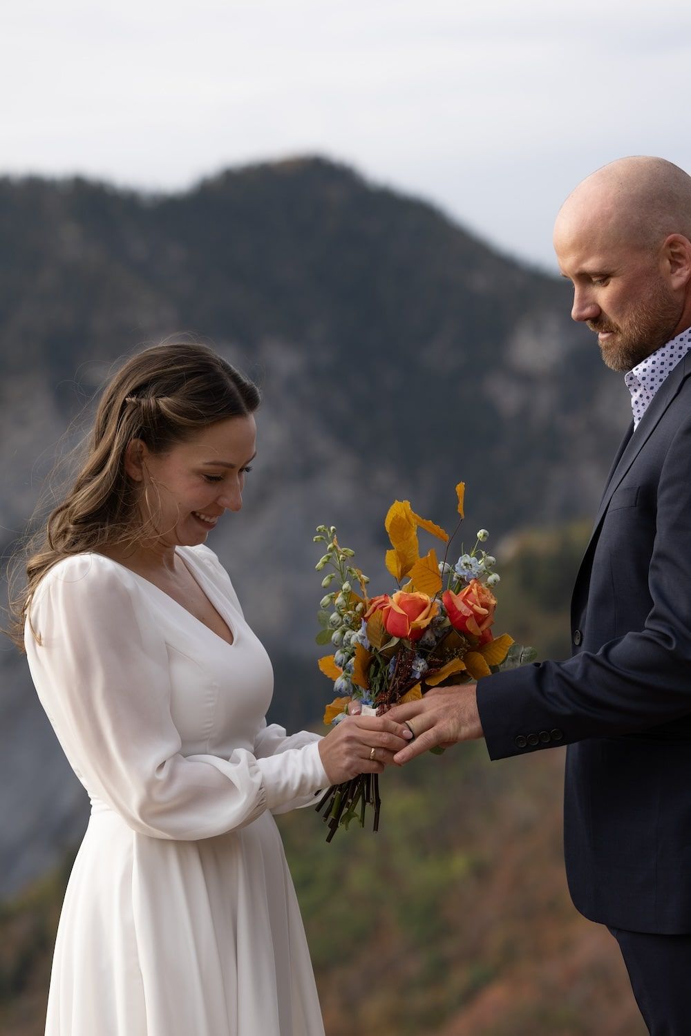 A couple shares a tender moment exchanging colorful flowers against a mountainous backdrop during their wedding ceremony.