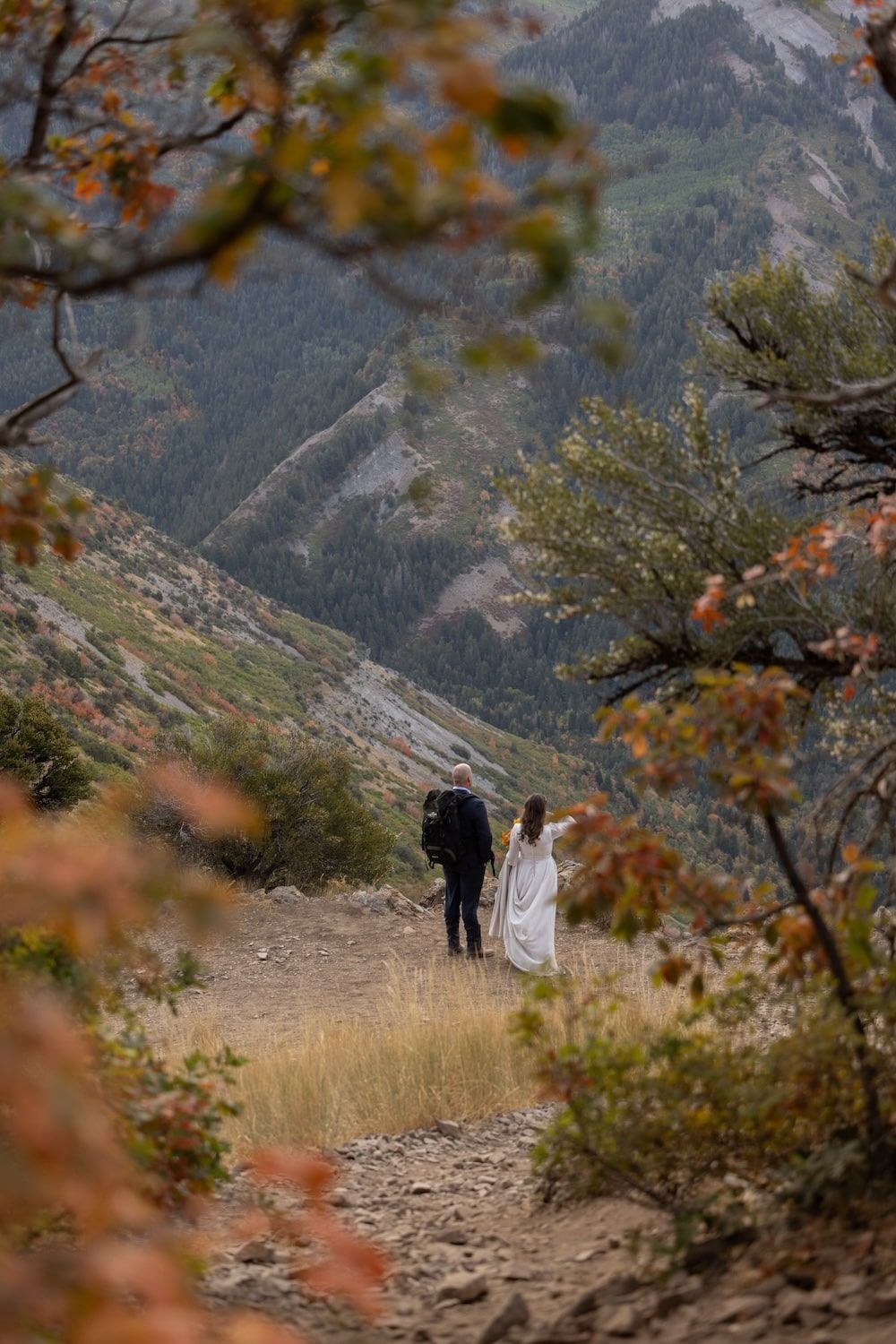 A couple in wedding attire stands on a rocky mountain trail surrounded by autumn foliage and scenic mountain views.