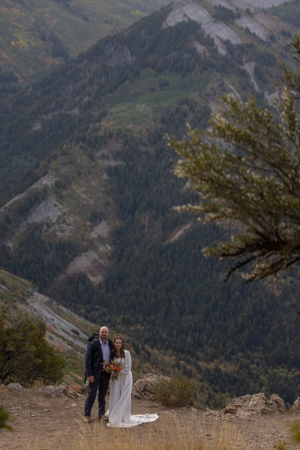 A couple stands together on a mountain overlook with dramatic mountain peaks and autumn foliage in the background.