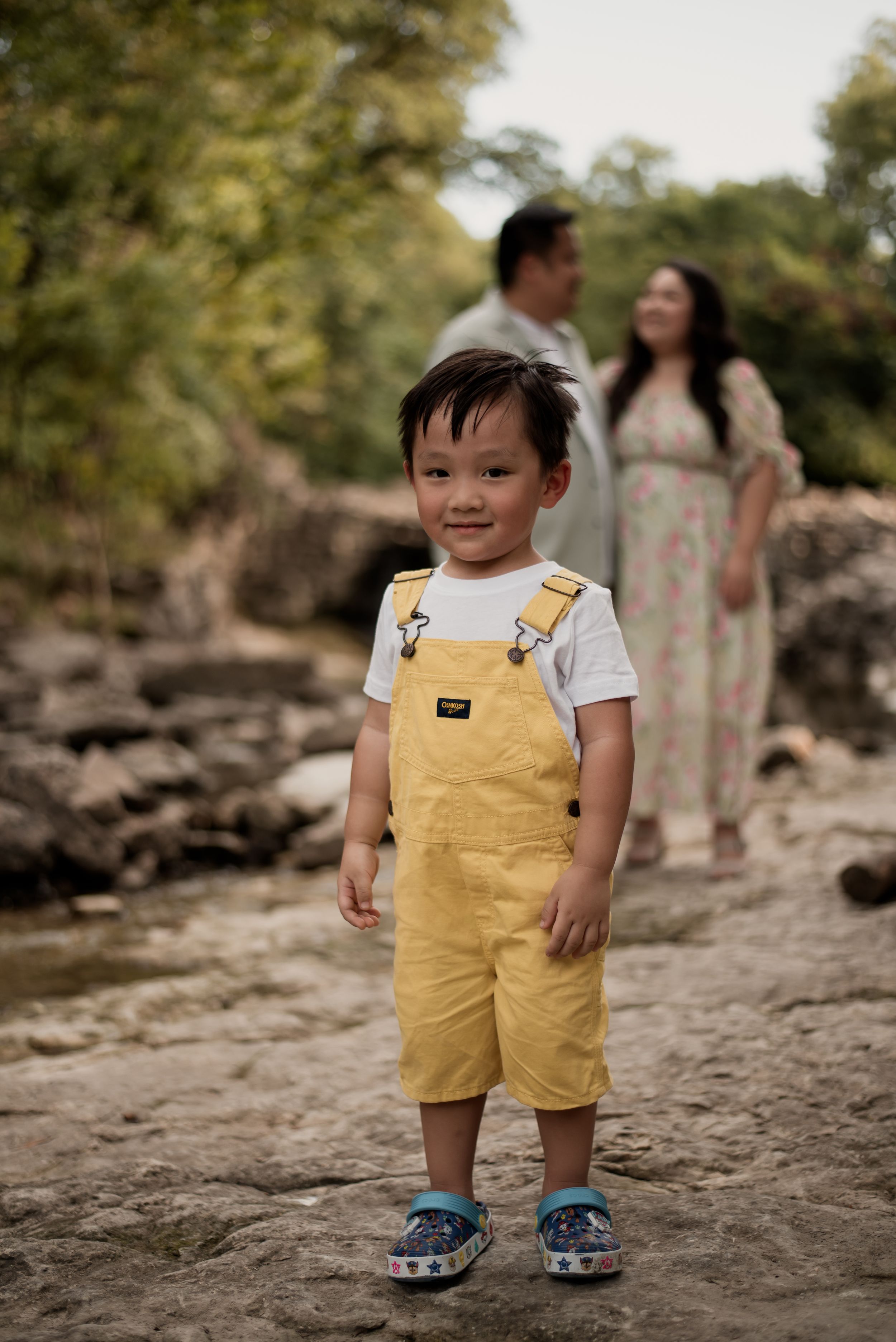 Toddler boy in yellow overalls smiles outdoors with parents standing behind him near rocky terrain.