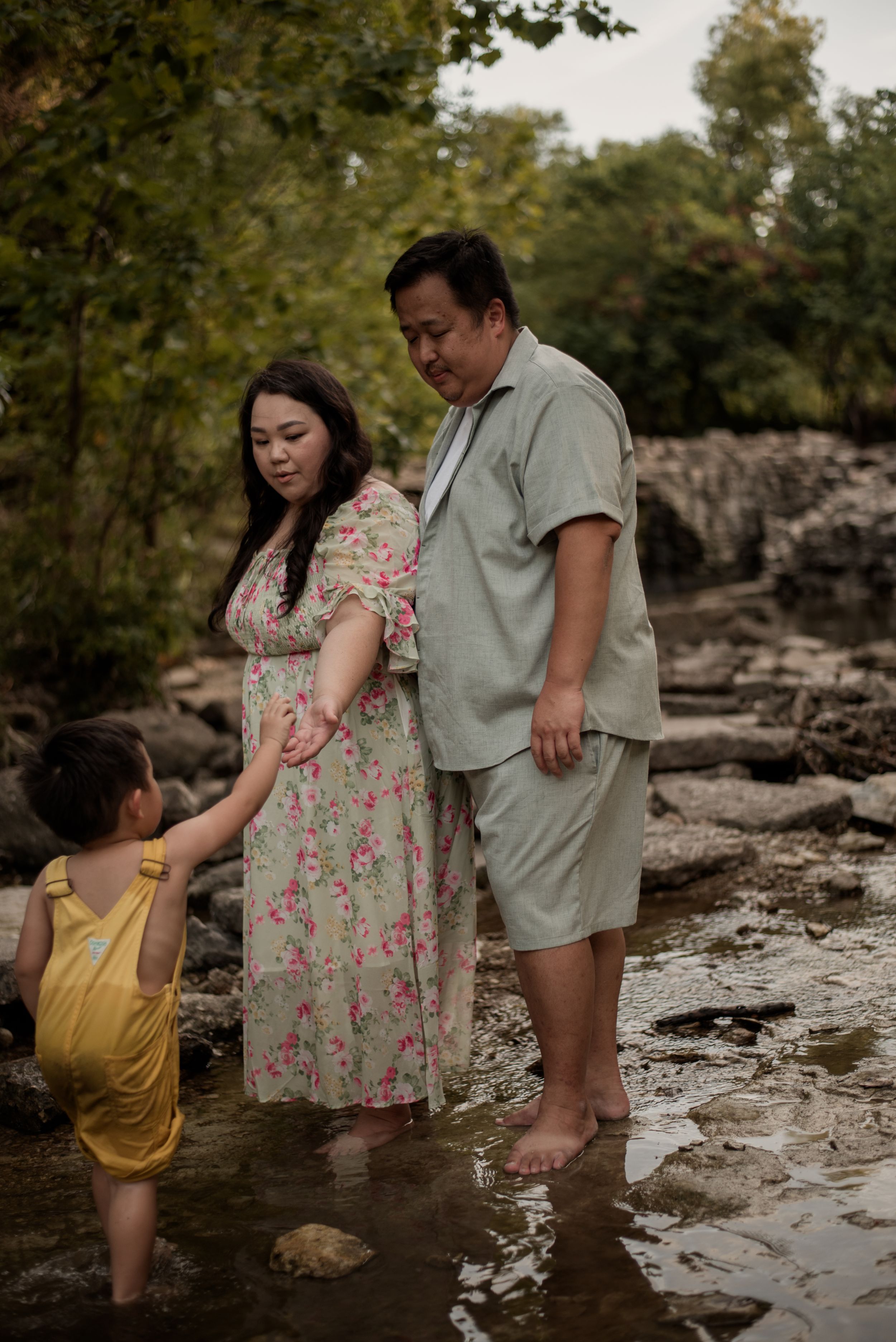 Asian family of three standing in a shallow creek, parents watching young child in yellow romper play in water.
