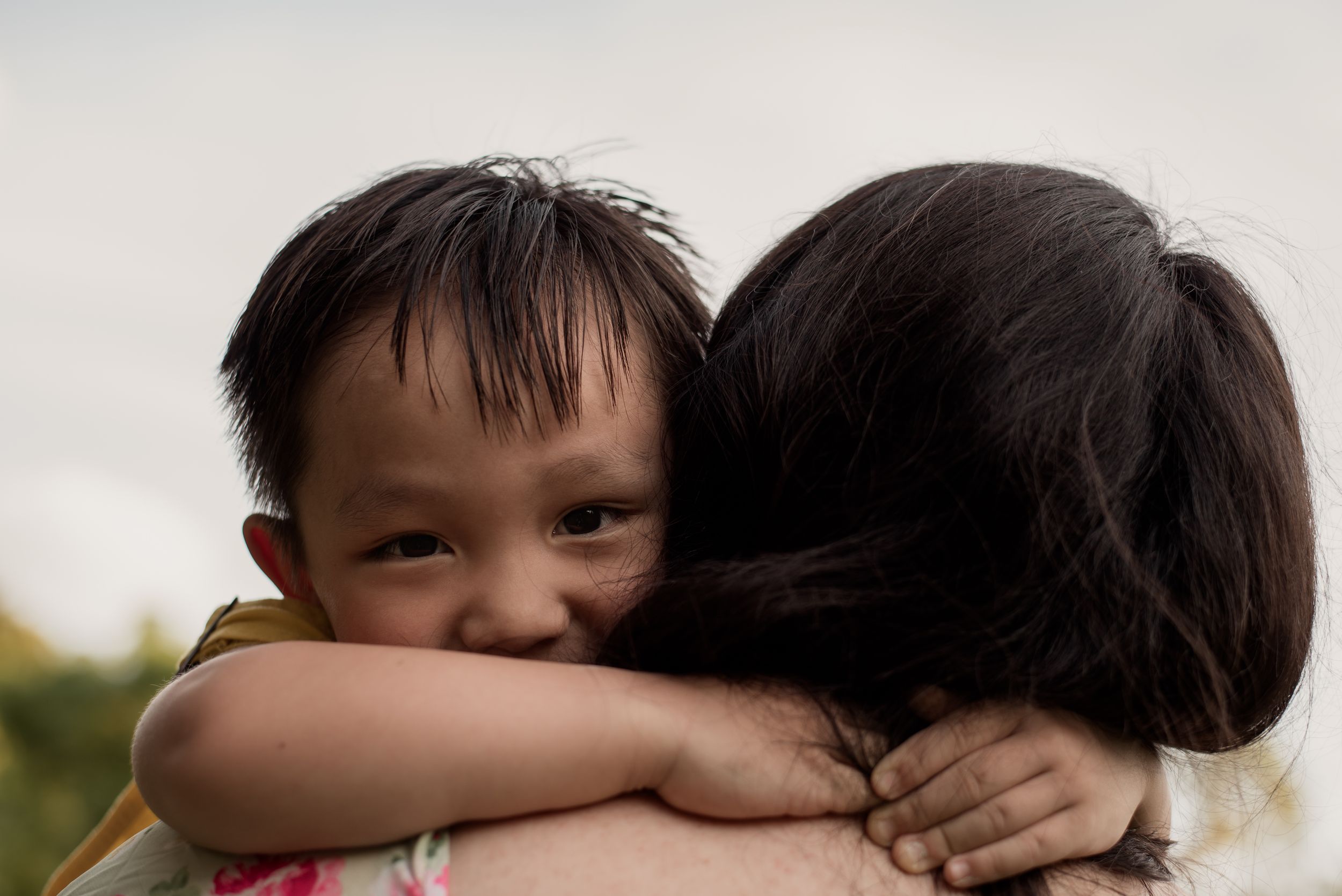 A young Asian child hugs a woman tightly, looking over her shoulder with a gentle smile outdoors.