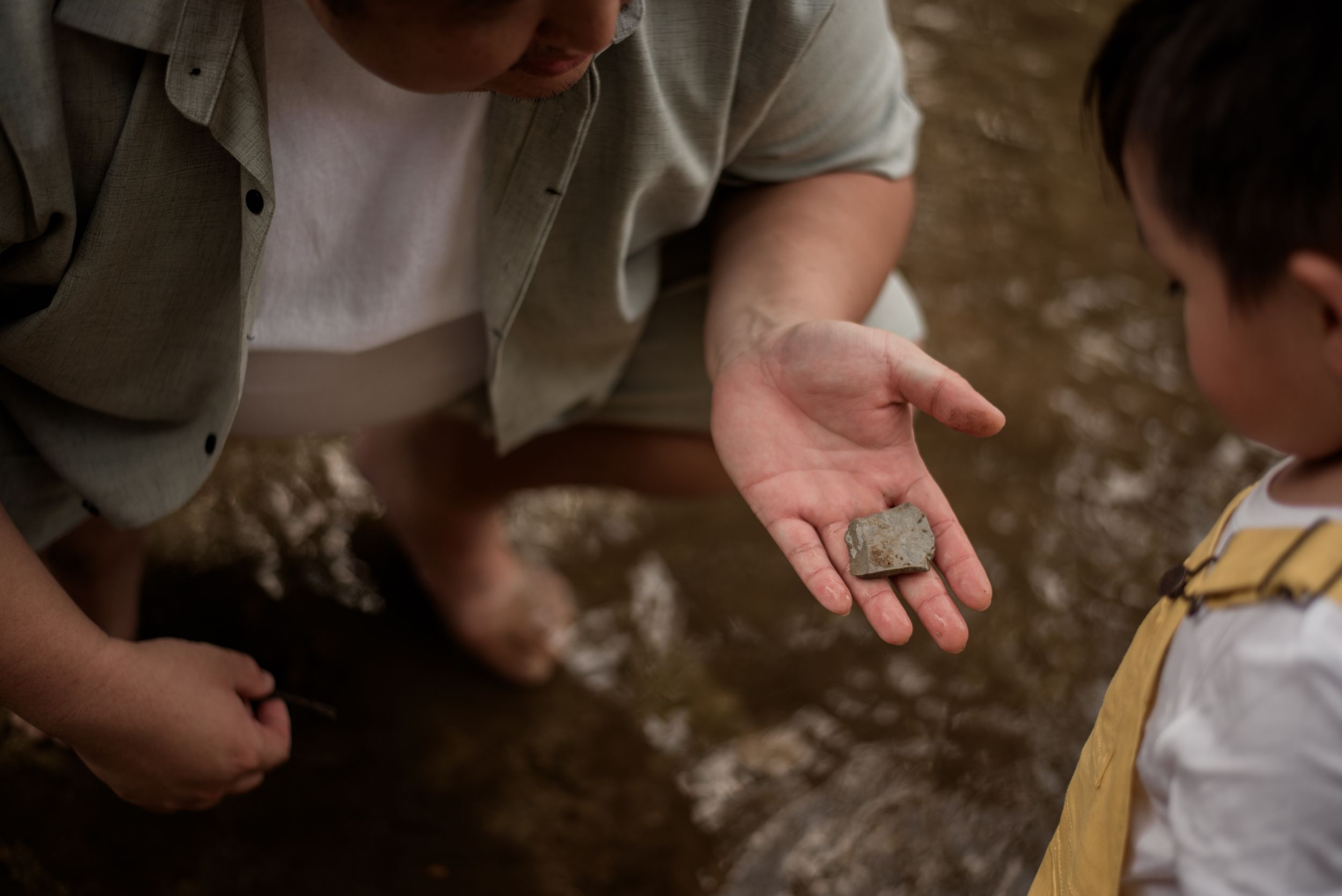 Child showing a small found rock or coin in an open palm to another child near a shallow stream outdoors.