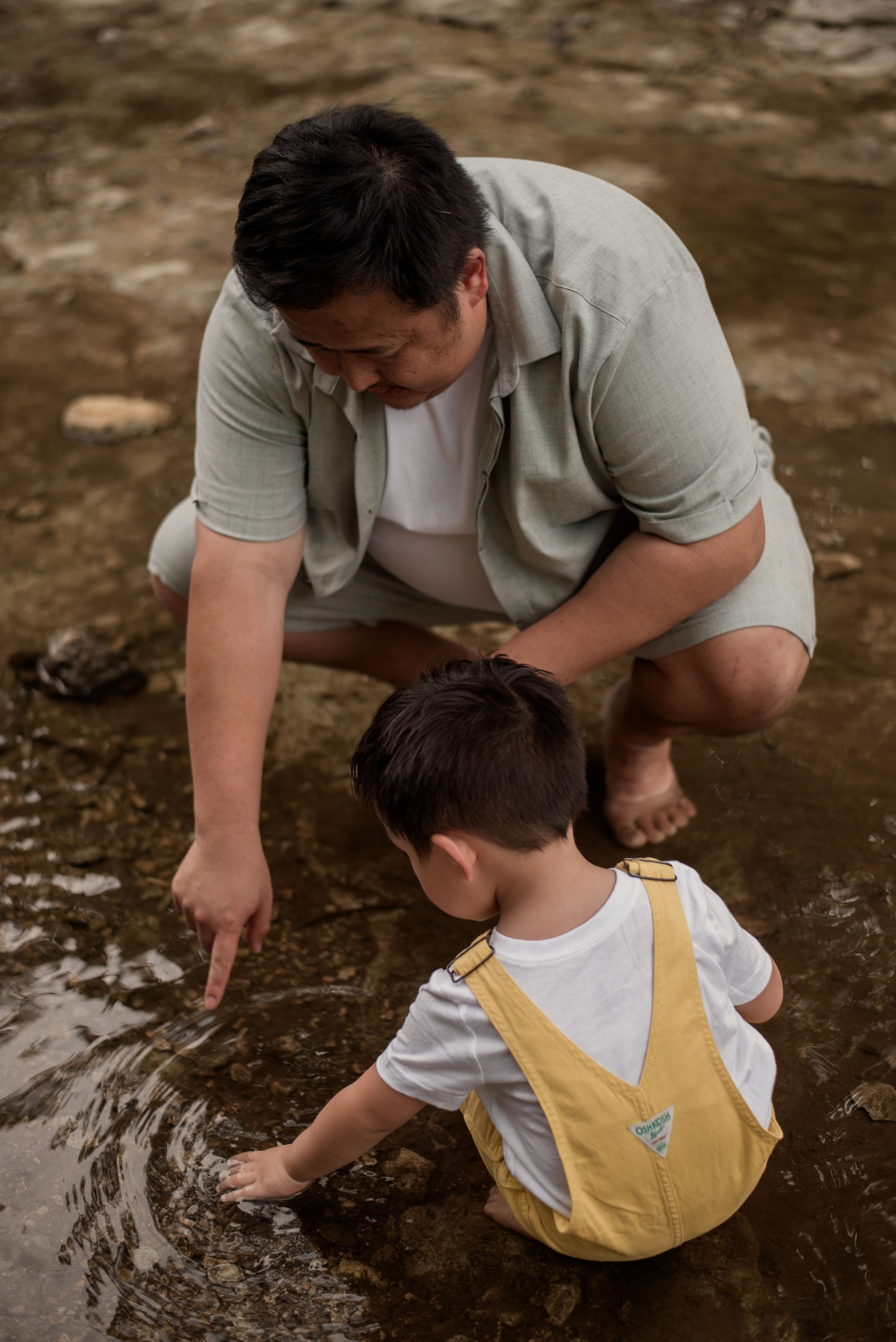 A father crouches beside his toddler son in yellow overalls, both exploring shallow water together outdoors.
