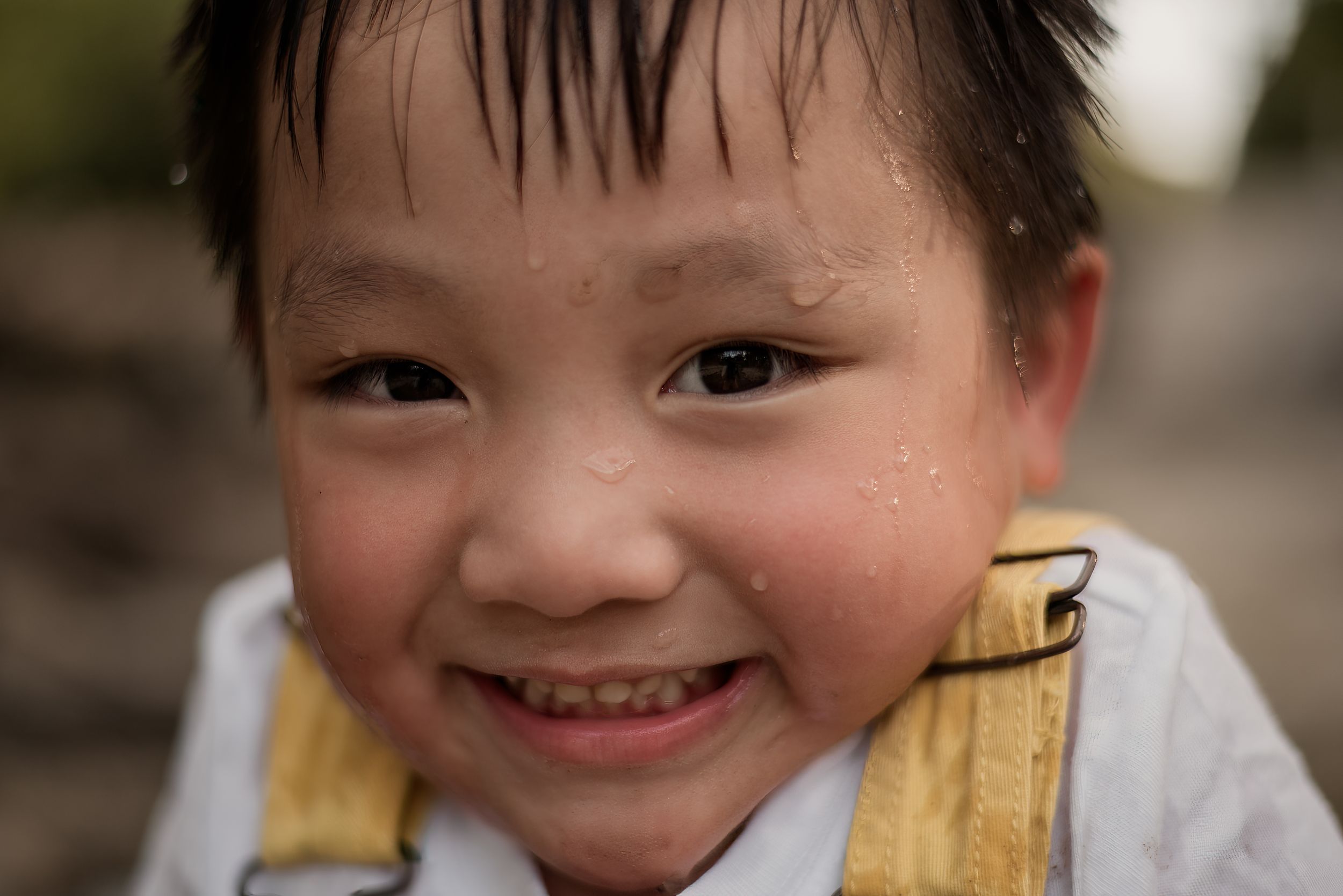 Smiling young Asian boy with wet hair and water droplets on his face, wearing yellow suspenders outdoors.