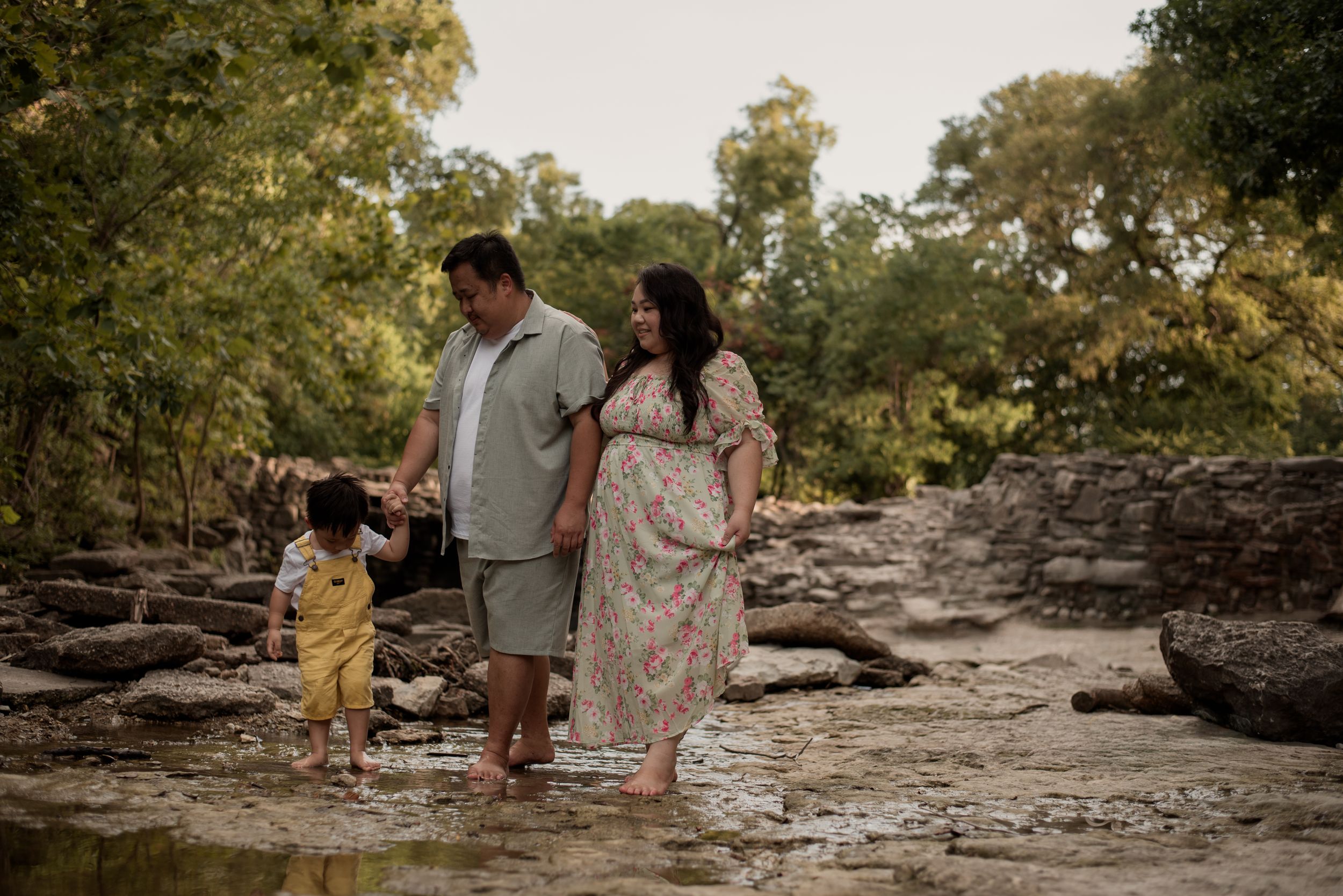 A pregnant woman in a floral dress walks with her partner and toddler along a rocky creek surrounded by lush trees.