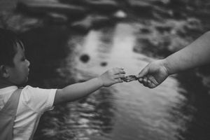 Black and white photo of a child reaching out to hold an adult's hand near a reflective body of water.