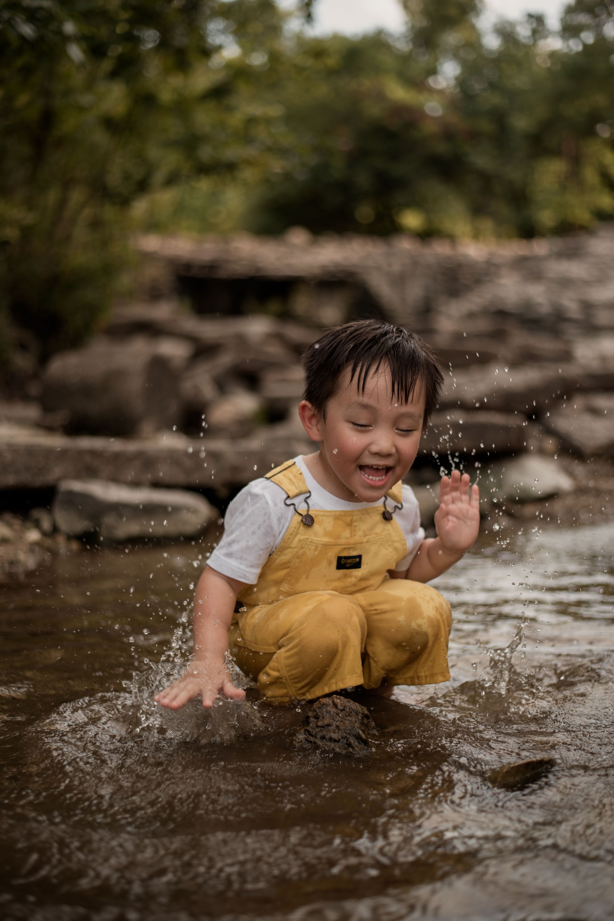 A joyful young boy in yellow overalls splashes and plays in a shallow rocky creek outdoors.