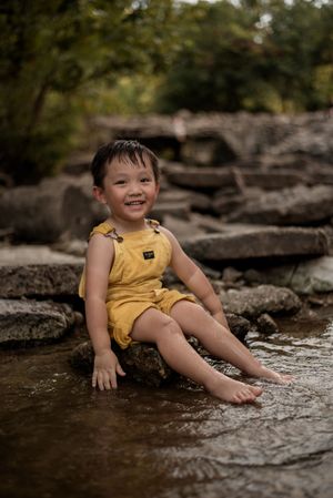 Smiling toddler in yellow romper sitting on rocks in a shallow creek surrounded by lush greenery.