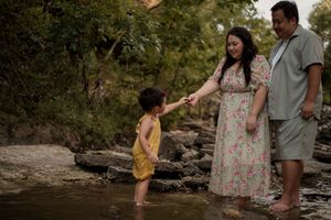 Family of three wading in a rocky creek, mother in floral dress holds child's hand while father watches.