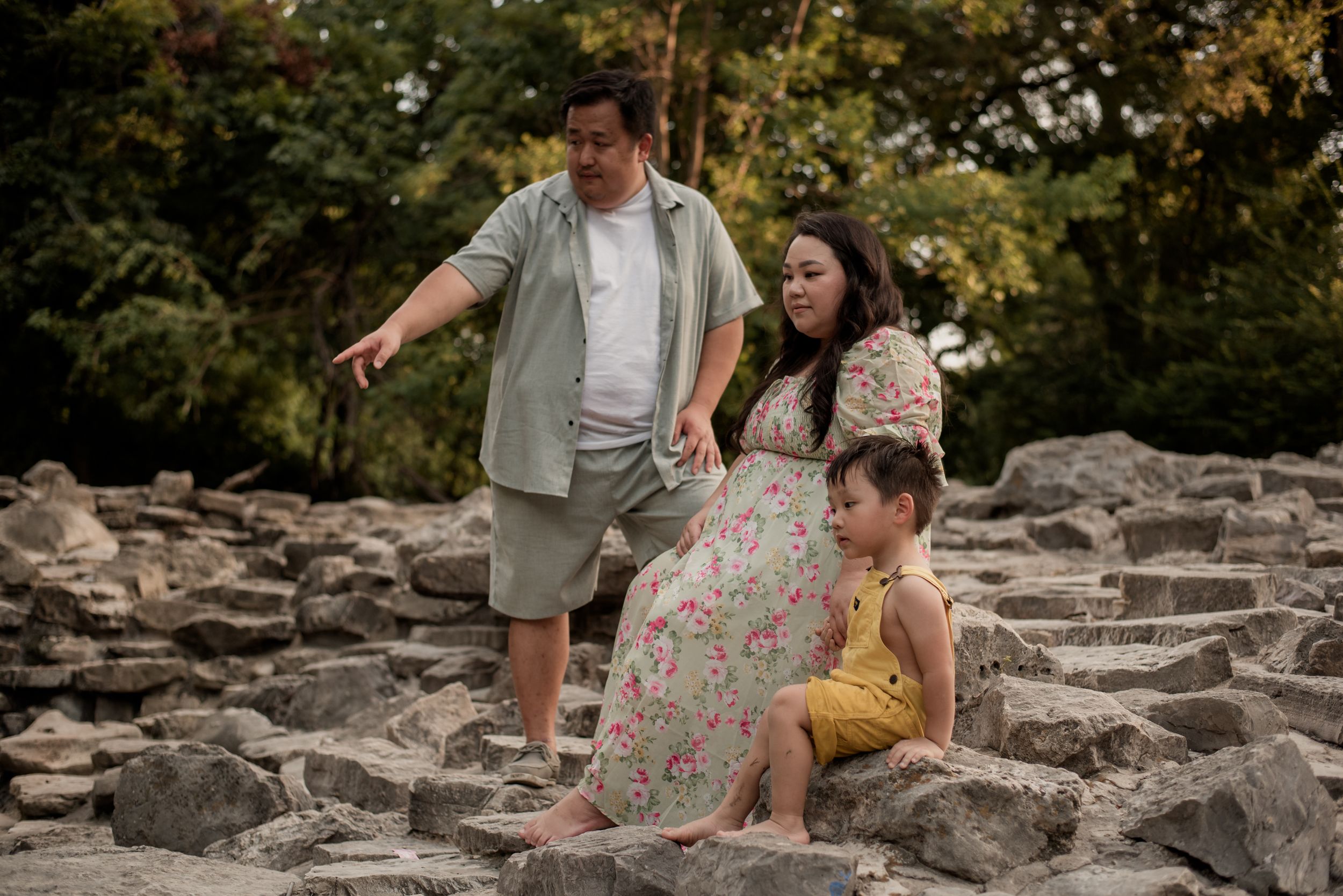 Family of three on rocky terrain, parents watching young child sit on stones, lush green trees in background.