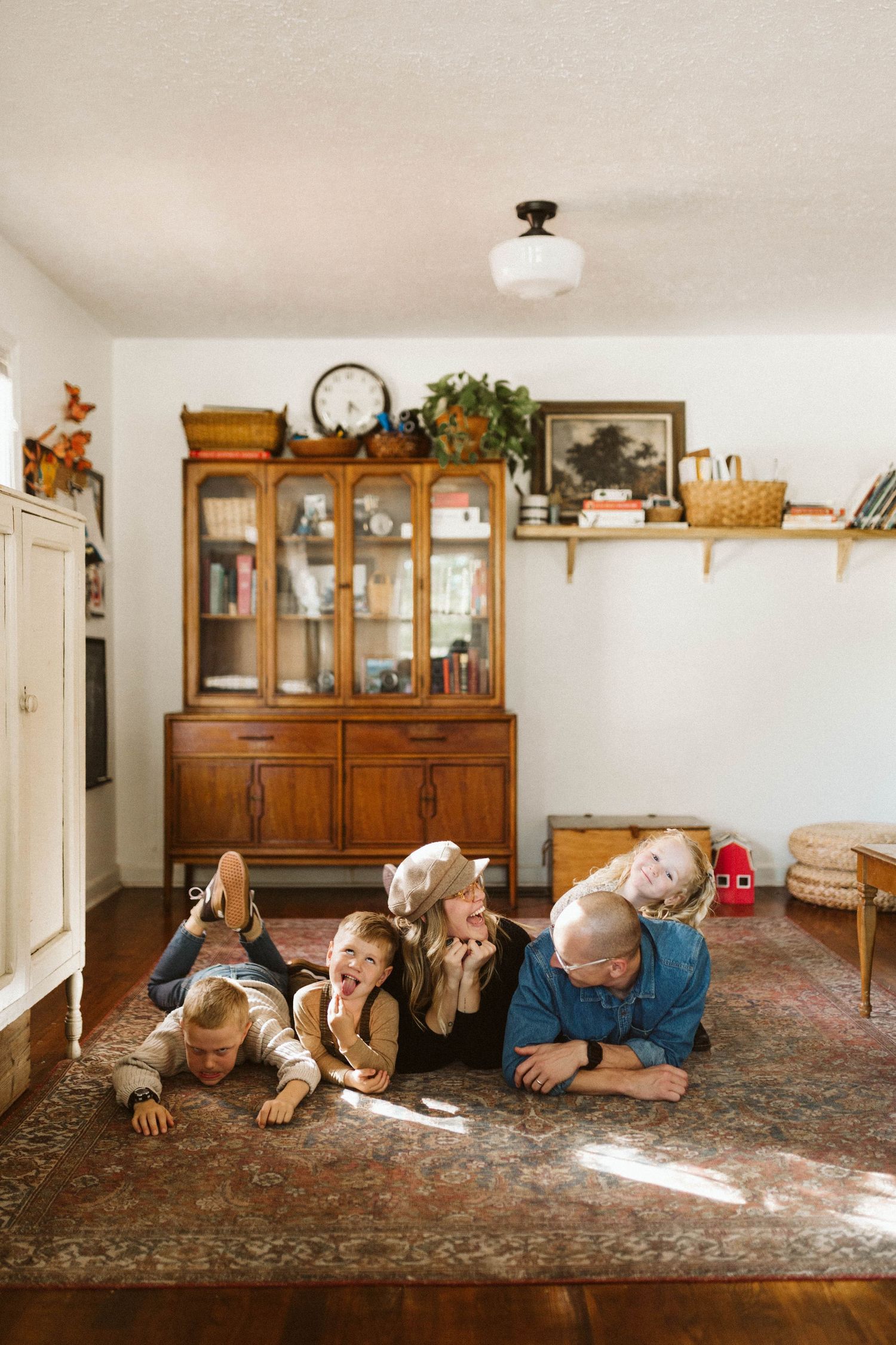 A family lies on a rug in a sunny room with vintage wooden furniture and decor in the background for a photoshoot with B. Logsdon.