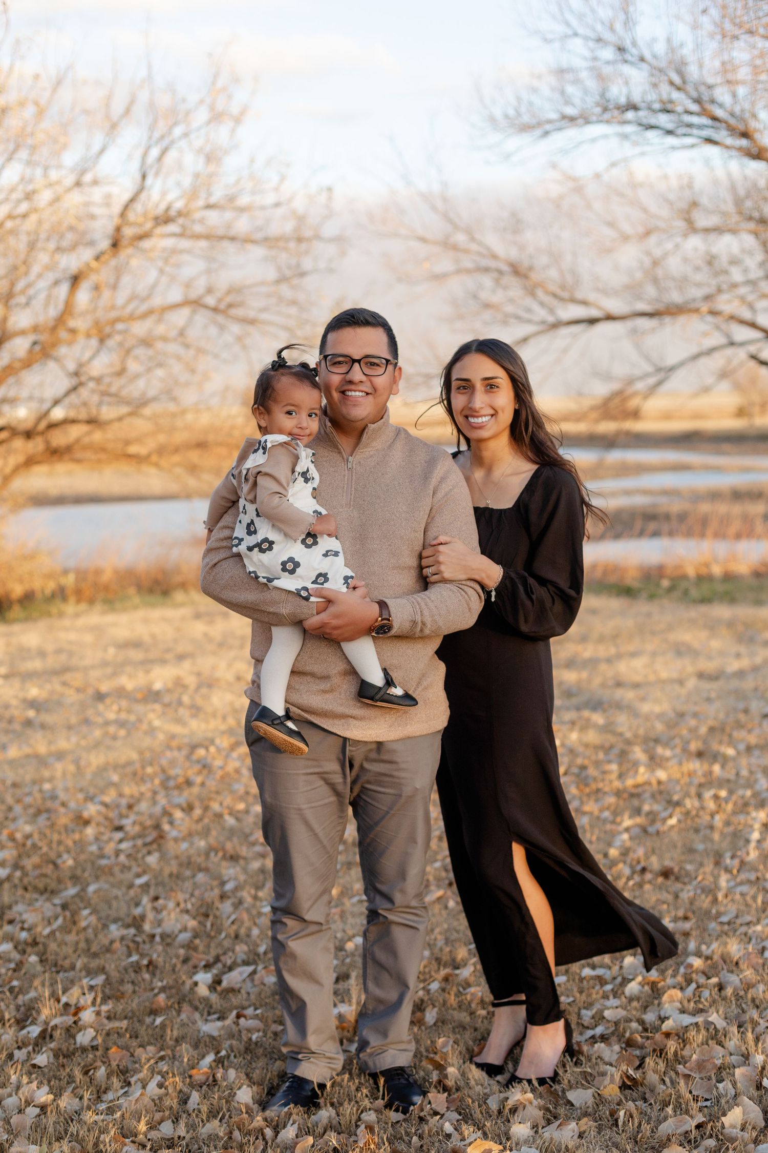 A family portrait taken outdoors during autumn with a river and bare trees in the background.