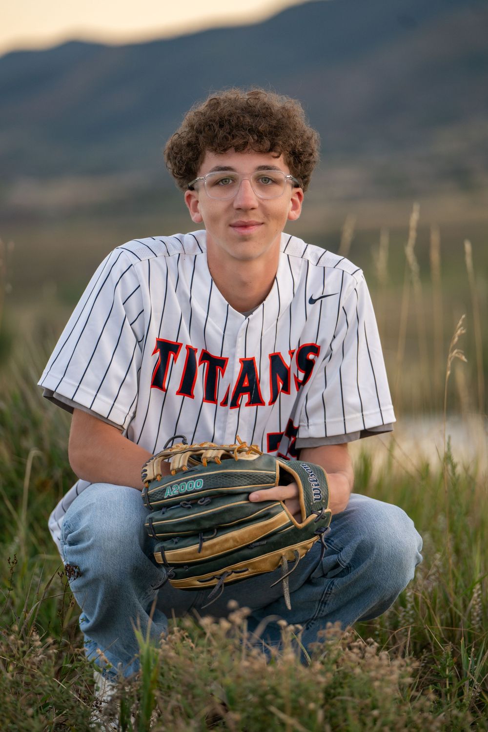 Baseball player in white pinstriped Titans jersey kneels in grass field at sunset holding glove.