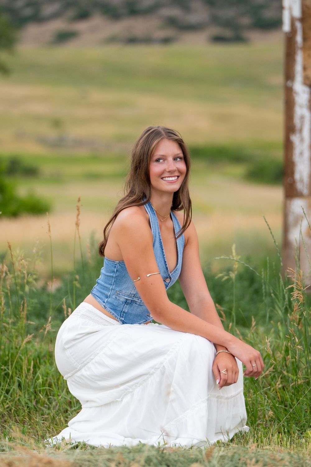Woman in denim vest and white maxi skirt poses in grassy field with rolling hills in background.
