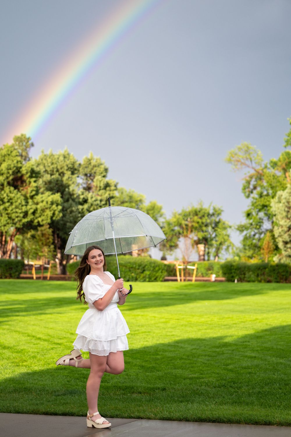 A whimsical portrait with a clear umbrella and rainbow in a green park setting. High school senior.