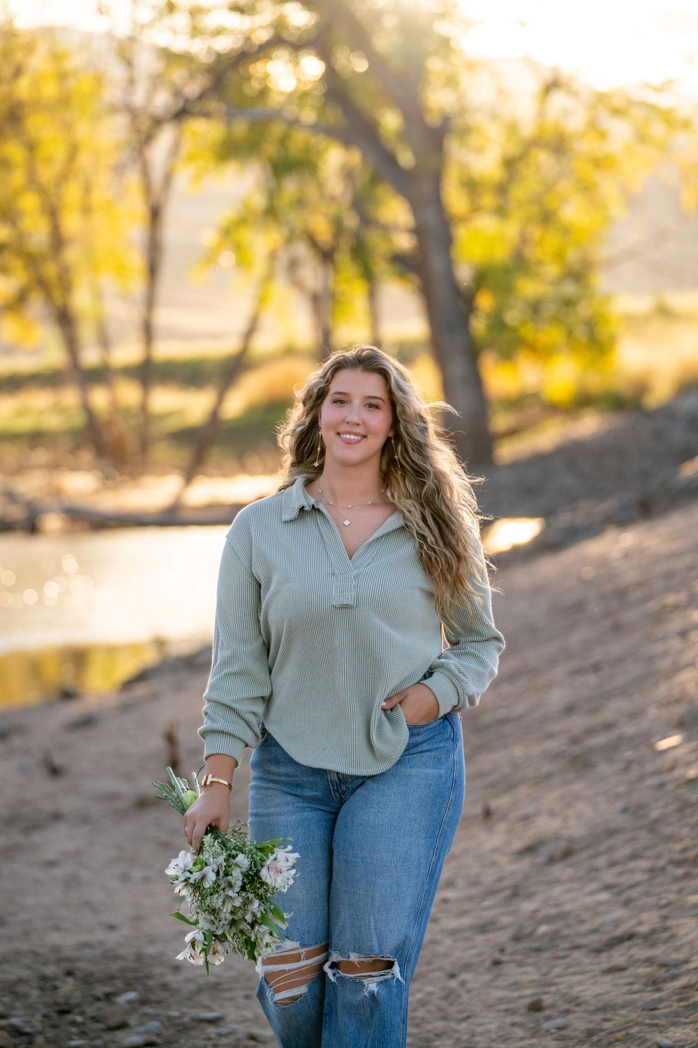 A person holding wildflowers poses in a green sweater and ripped jeans during golden hour. High school senior.
