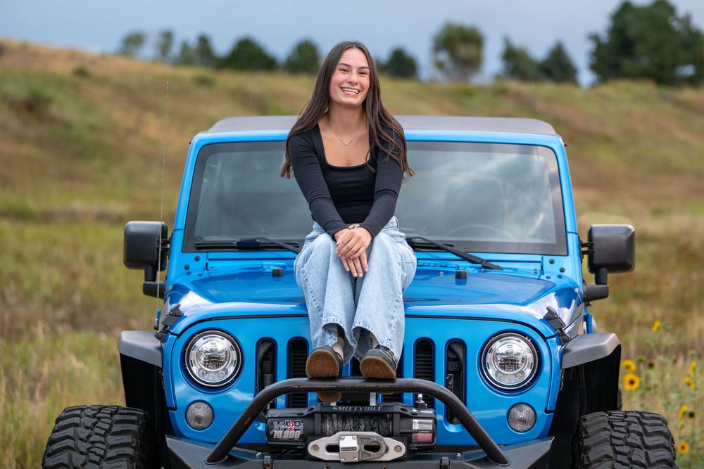 Someone sitting on the hood of a bright blue Jeep Wrangler in an open field. High school senior.
