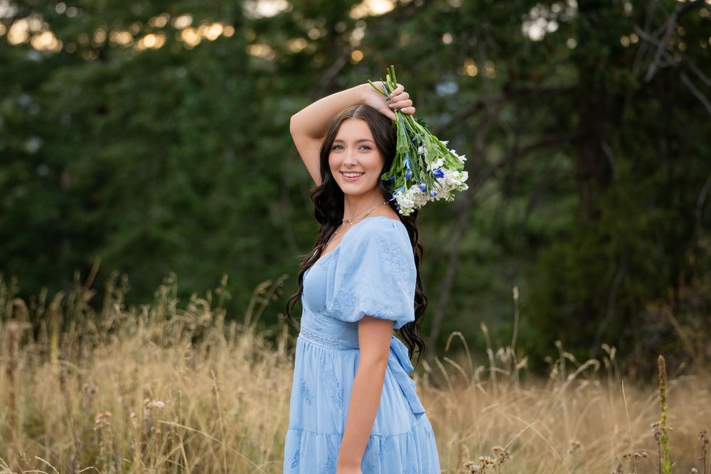 Woman in light blue summer dress holds wildflower bouquet while standing in golden field at sunset.