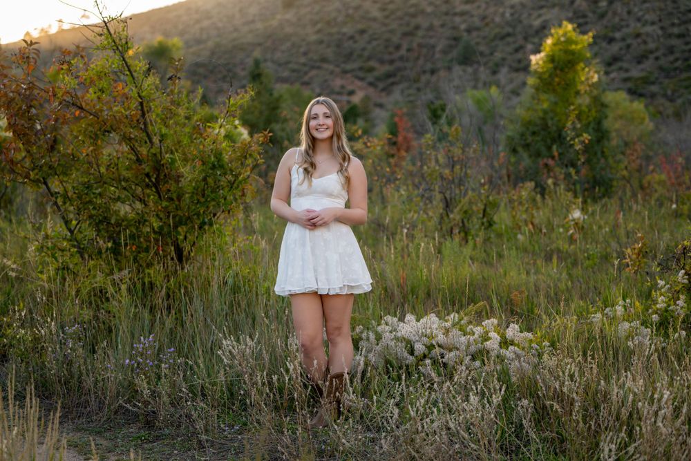 Woman in white dress stands in tall grass with mountains in background at sunset. High school senior.