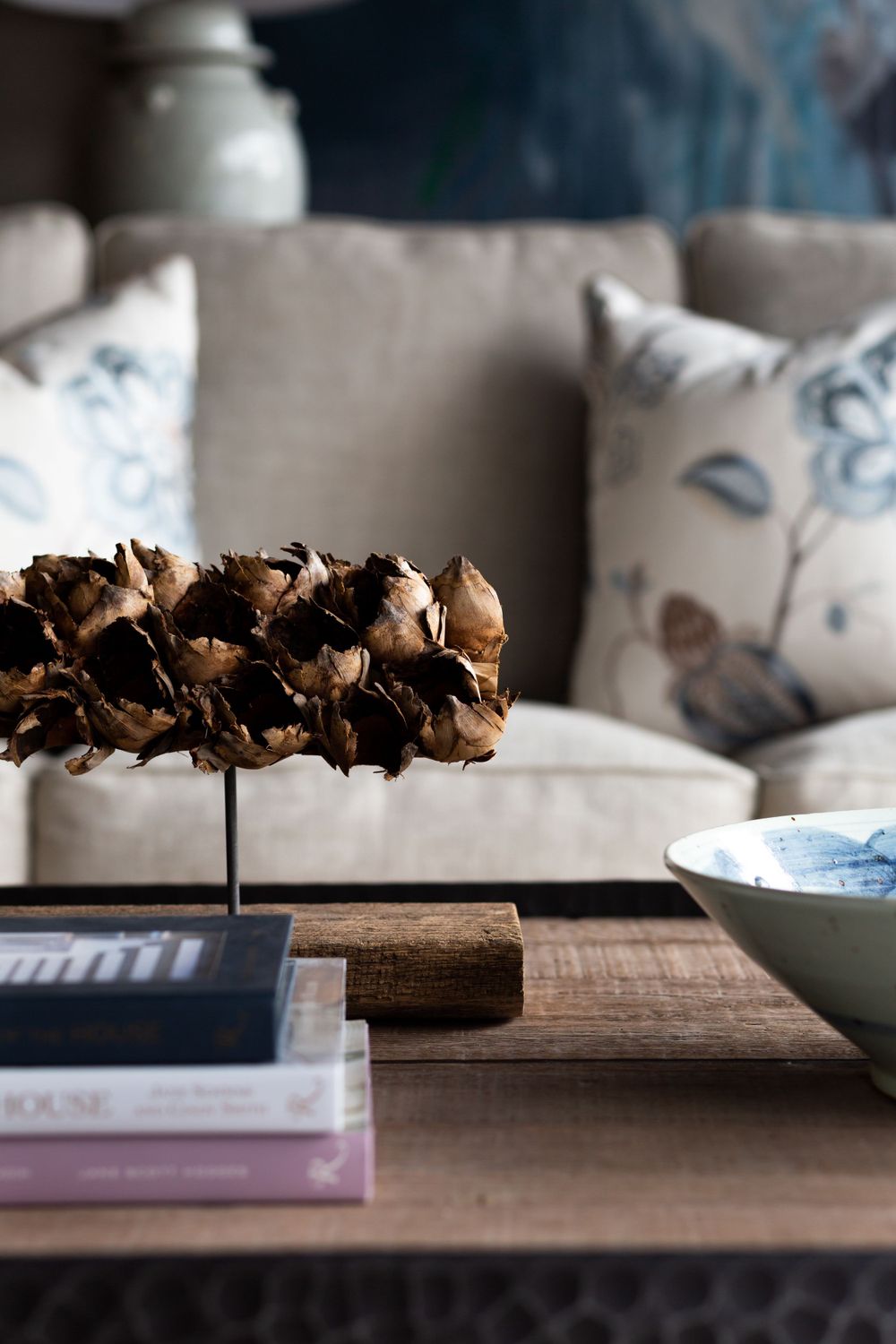 Dried flowers displayed on stand with books and pillows on couch in cozy living room setting.