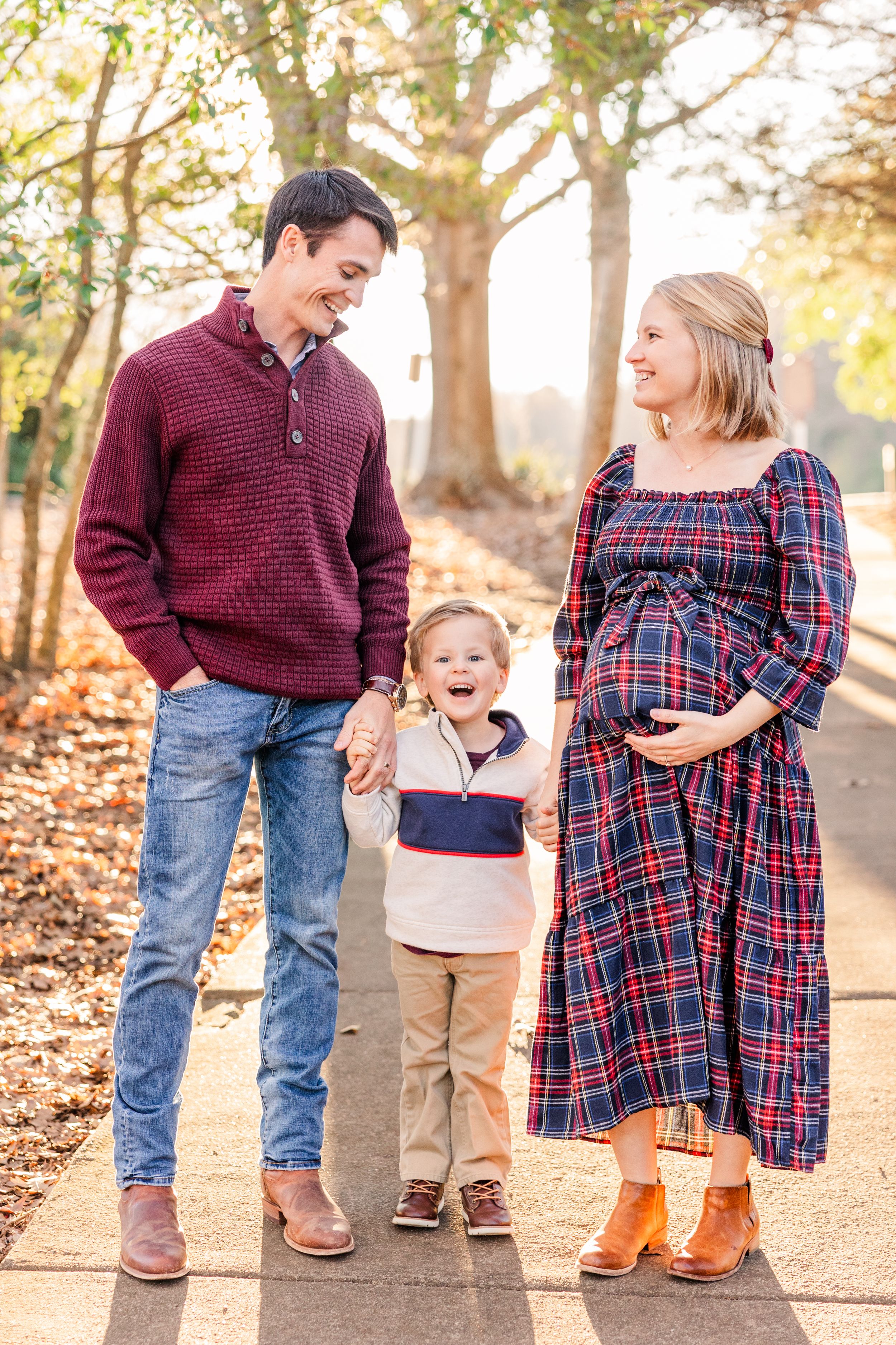 Family standing together outdoors with expectant mother in plaid dress and child holding parents' hands.