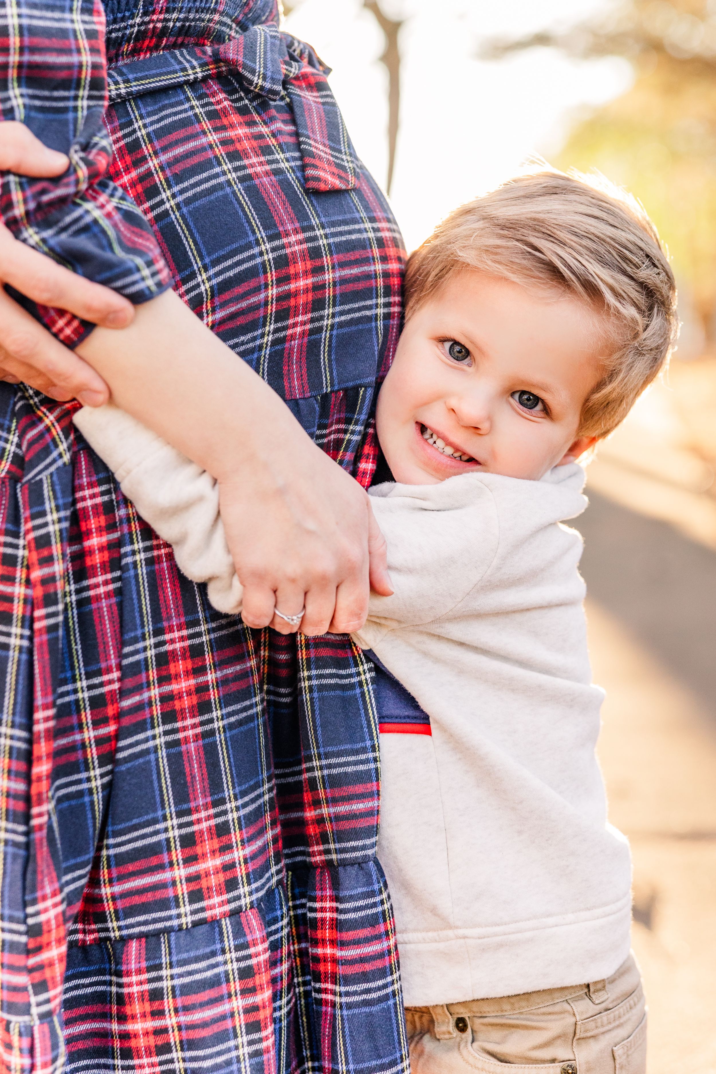 Young child hugging pregnant mother wearing plaid dress during outdoor photo session.