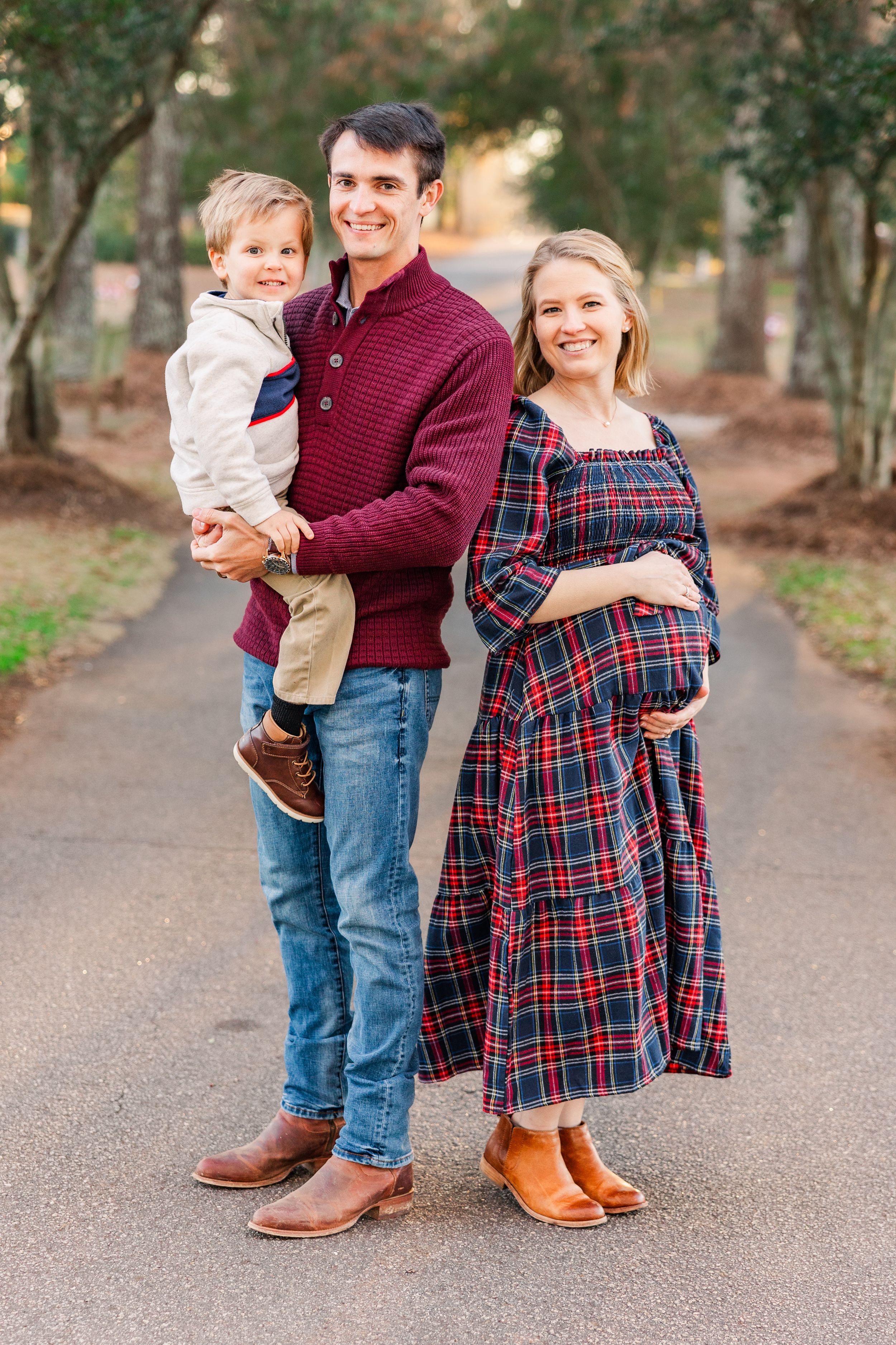 Family portrait with father in burgundy sweater, pregnant mother in plaid dress, and young child.