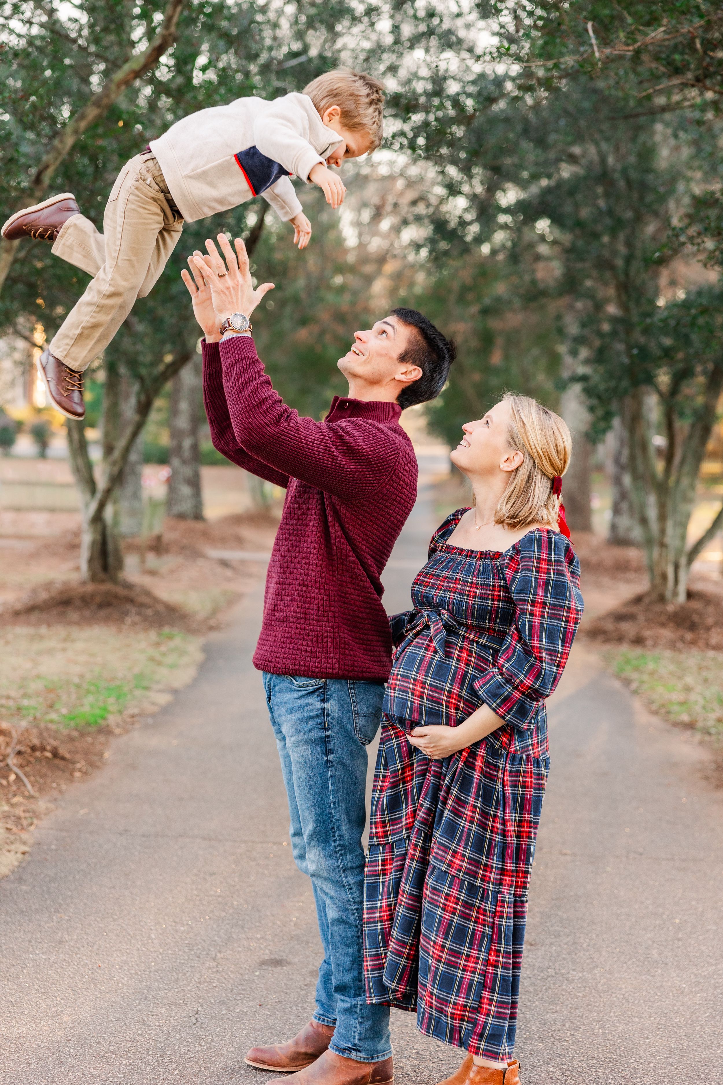 Father lifts child in air while pregnant mother in plaid dress watches during outdoor photoshoot.