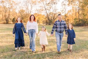 Family holds hands while walking through a sunlit field wearing coordinated blue and white outfits.