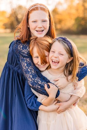 Three children in blue and cream dresses share joyful laughter during outdoor portrait session.