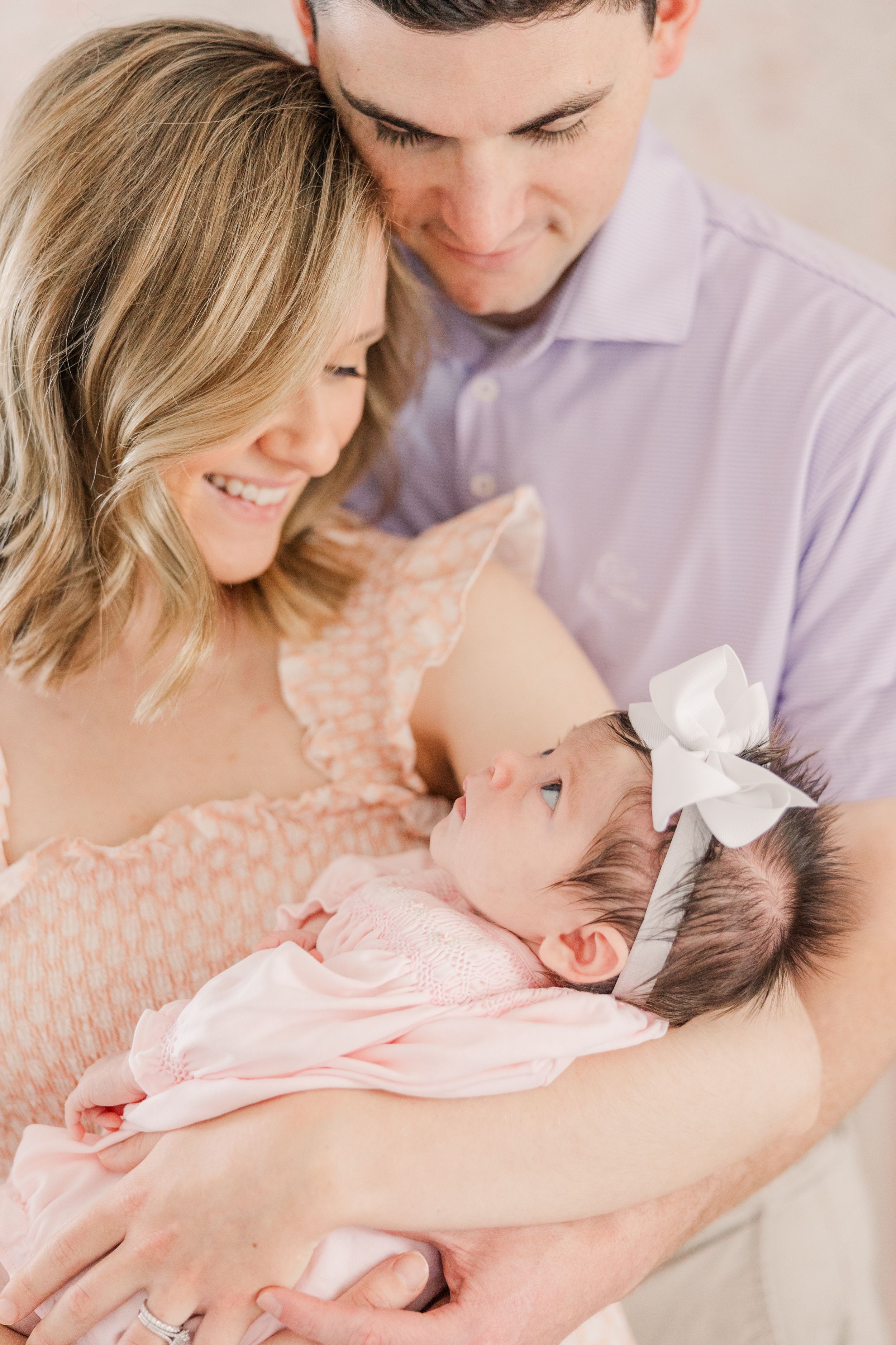 Parents smiling adoringly at their newborn baby wearing a white bow headband in a tender family moment.