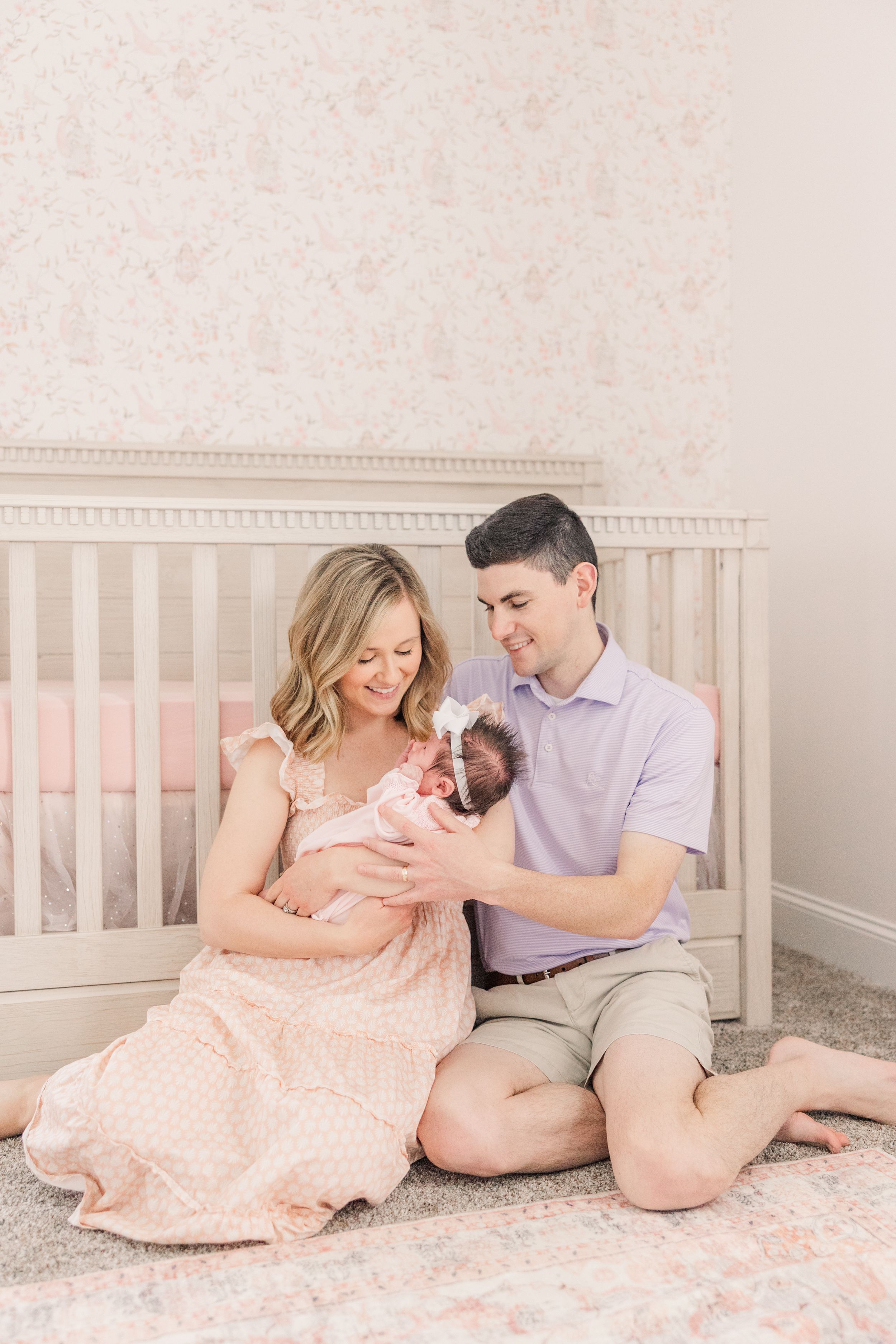 New parents sitting on floor by white crib sharing intimate moment with their baby in pink nursery.