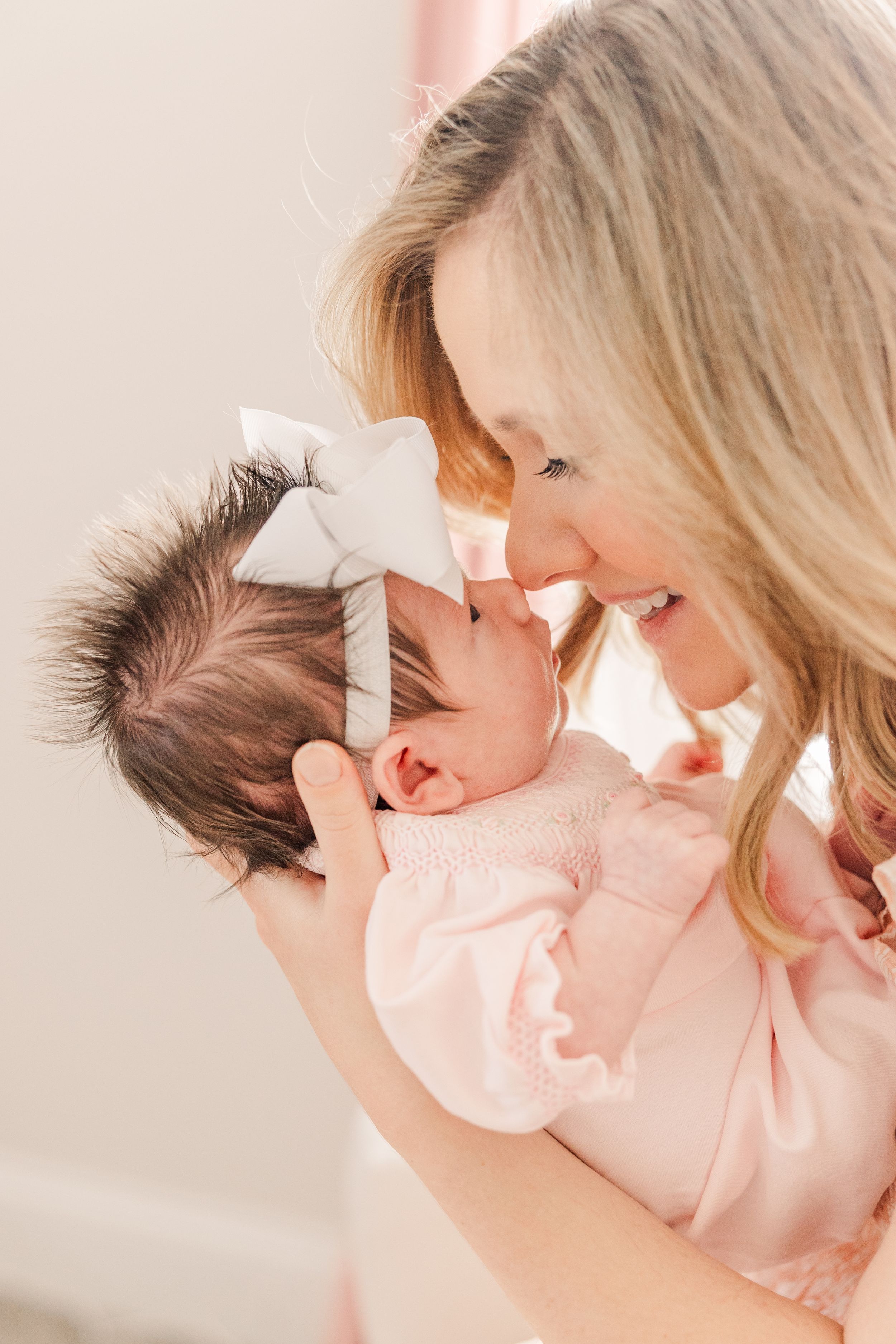 Mother in pink dress sharing tender nose-to-nose moment with newborn wearing white bow.