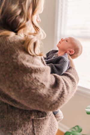 Baby in gray sweater being held up in natural window light.