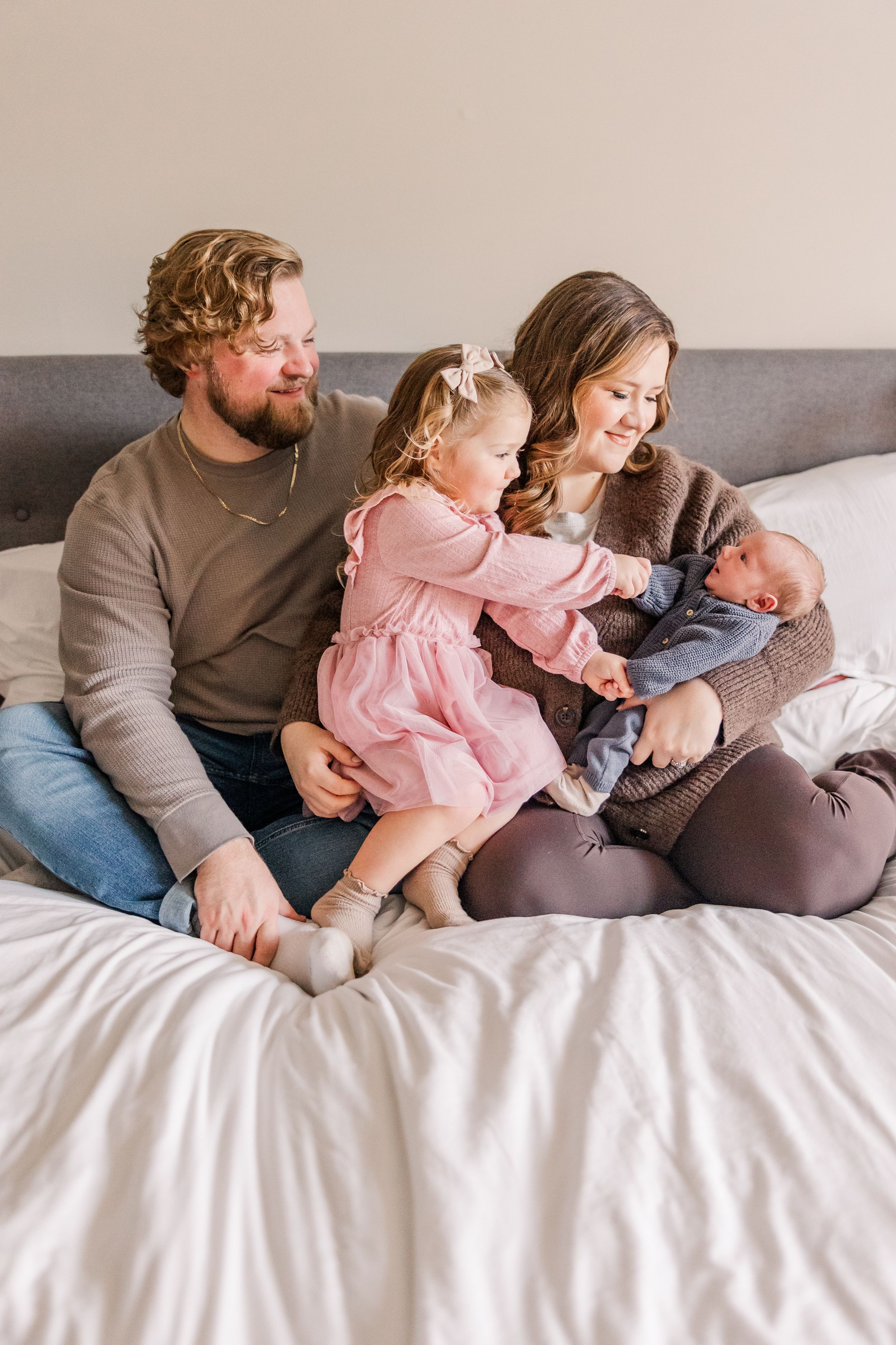 Family sitting together on bed with newborn baby and toddler in pink dress.