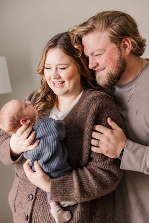 New parents share tender moment with their newborn baby while wearing cozy brown sweaters in intimate home portrait.