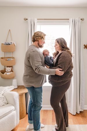 Parents holding newborn baby while standing by window in nursery with hanging storage baskets.
