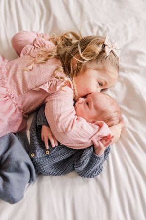 Siblings sharing a tender moment on white bedding as older sister in pink cuddles with baby wearing gray.