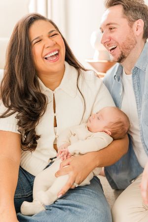 Candid family moment of laughter while holding sleeping baby in casual home setting.