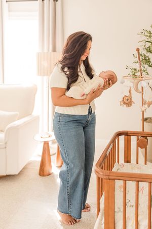 Parent holding baby in nursery with wooden crib and table lamp visible.