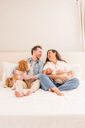 Happy family laughing together on white bed with dog and baby.