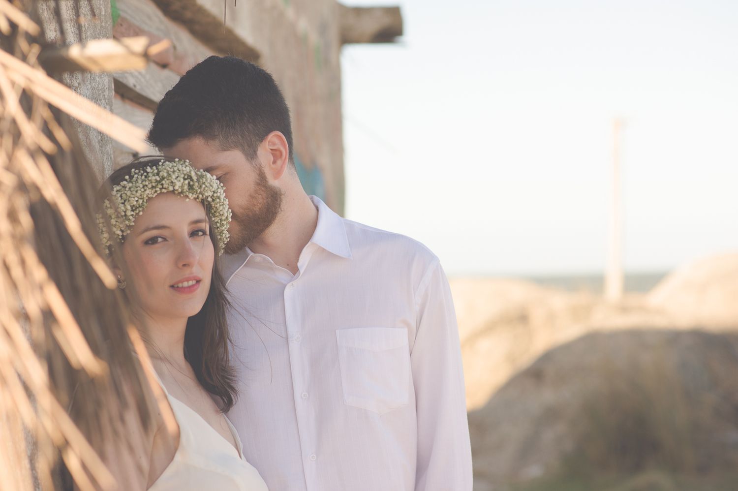 A romantic couple embraces during a beachside photoshoot at sunset, with her wearing a delicate floral crown.