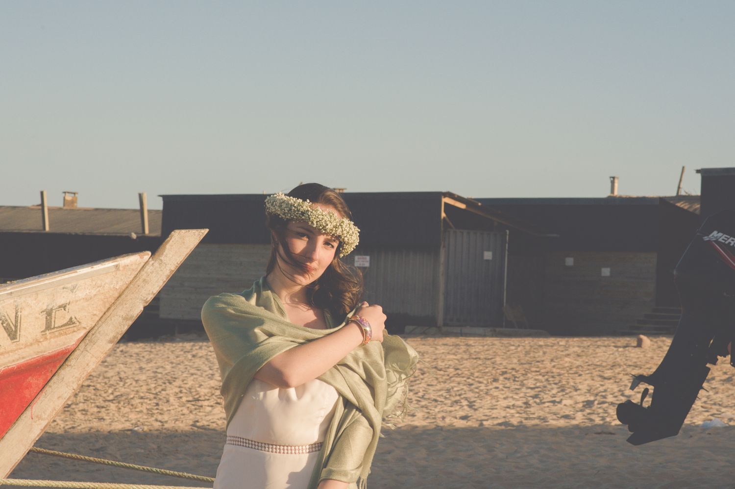 A wrapped figure stands in casual dress against a sandy beach backdrop.