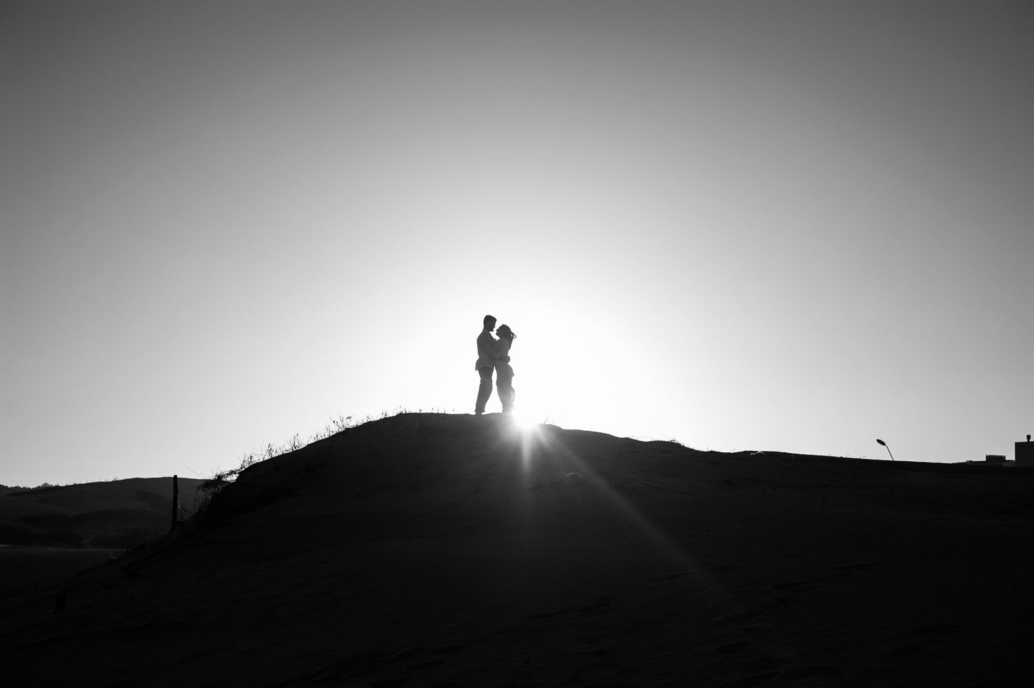 Two silhouettes stand together on a hilltop against a bright sunset sky.