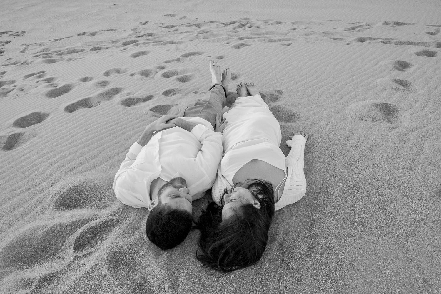 An overhead view of two people lying on sand dunes wearing white clothing.