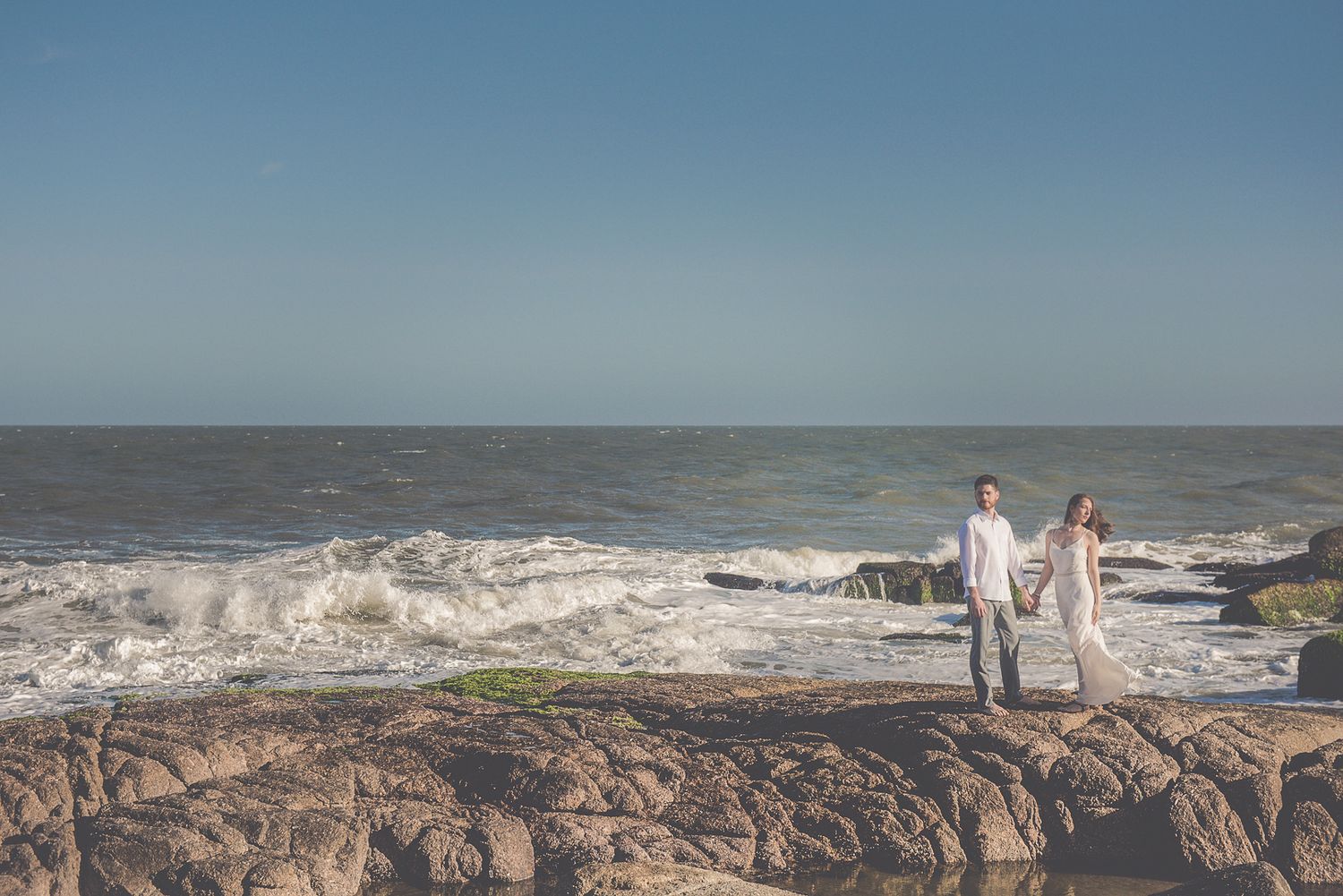 Couple holds hands while standing on rocky ocean shore under bright blue sky.